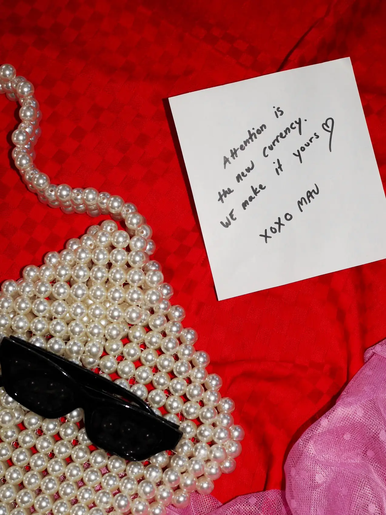 A white note with the handwritten text ‘Attention is the new currency. We make it yours. XOXO MAU’, placed on red fabric next to a pearl necklace.