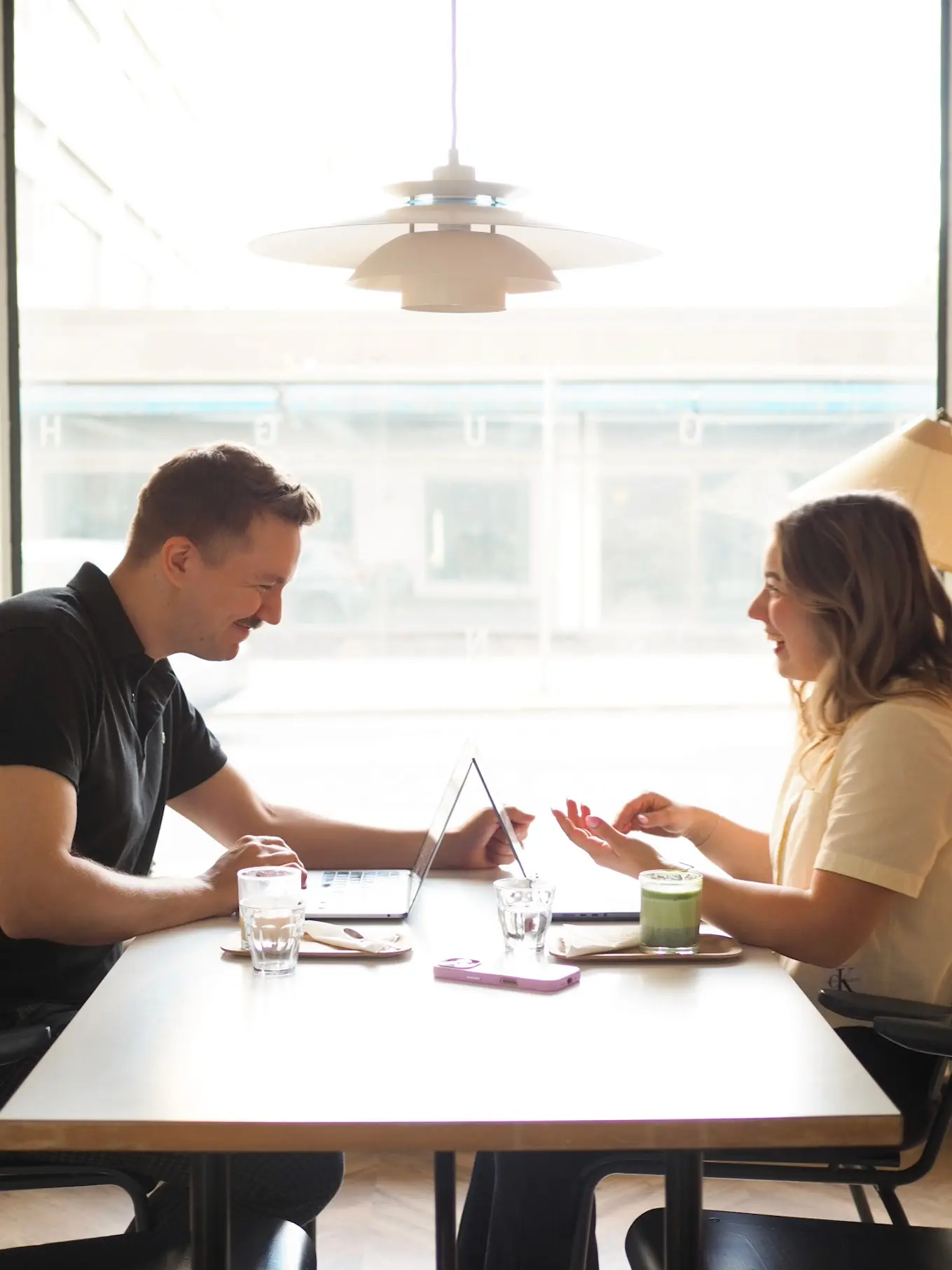 MAU Agency team members Saana and Totte sit at a cafe table, smiling and talking, with laptops, drinks and a lilac phone in front of them. They are planning a short-form video or working on content.