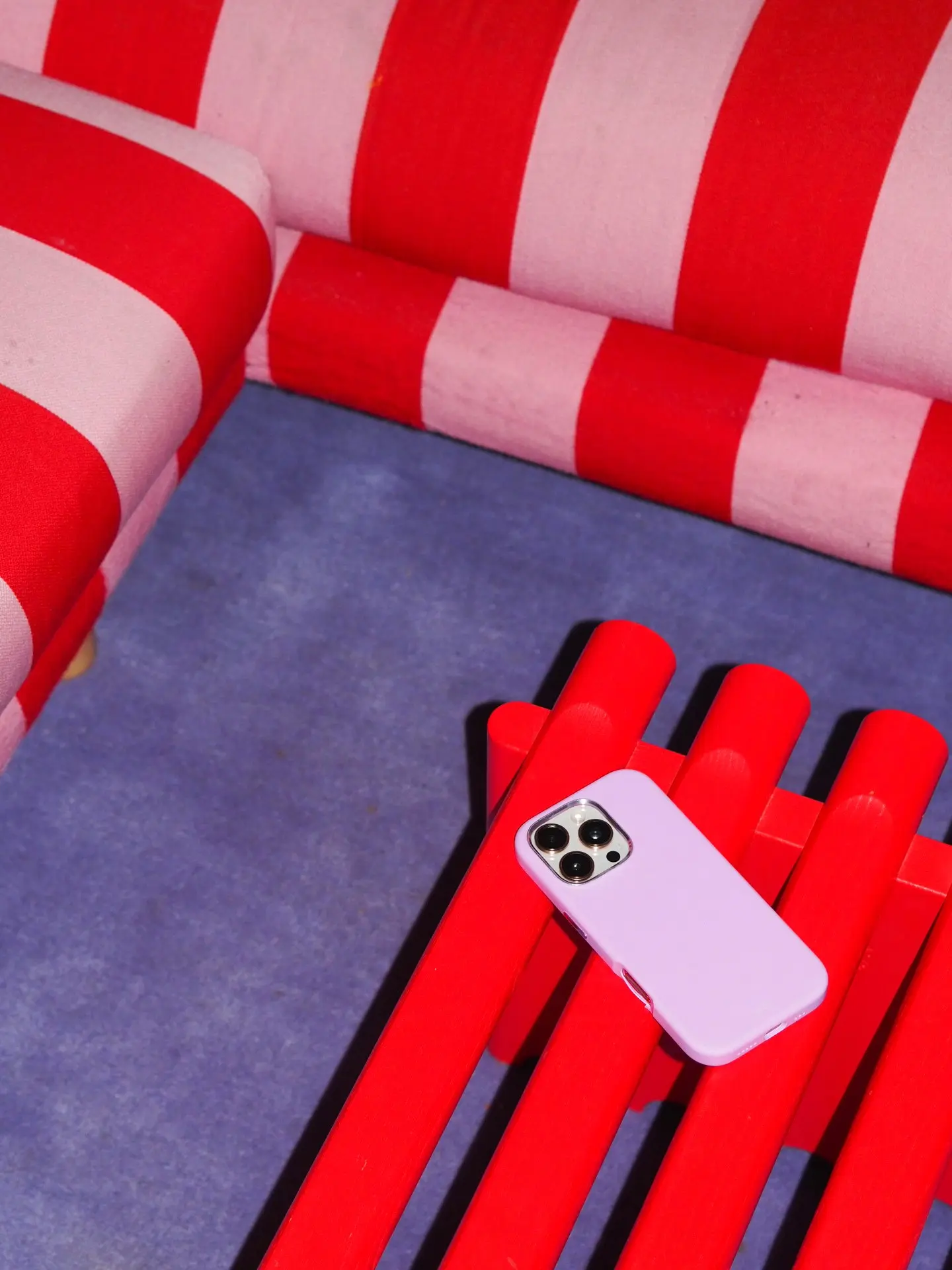 A lilac phone resting on a bright red bench on a blue floor, with red-and-white striped sofa cushions in the background.