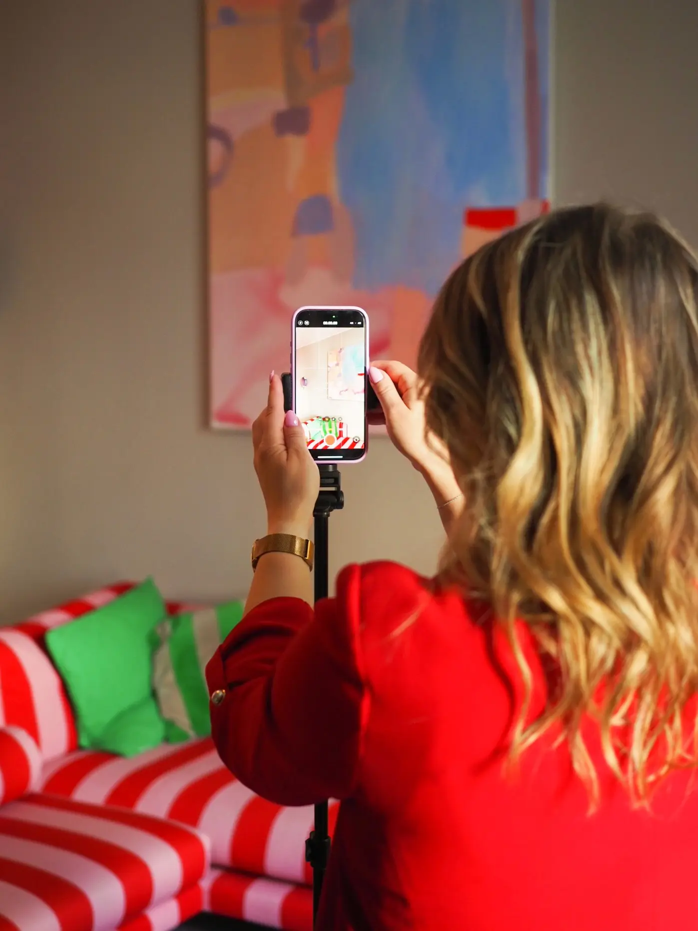 Saana from MAU Agency films a short-form video on her phone, mounted on a tripod. She stands in a colorful interior with a red-and-white striped sofa and green cushions.
