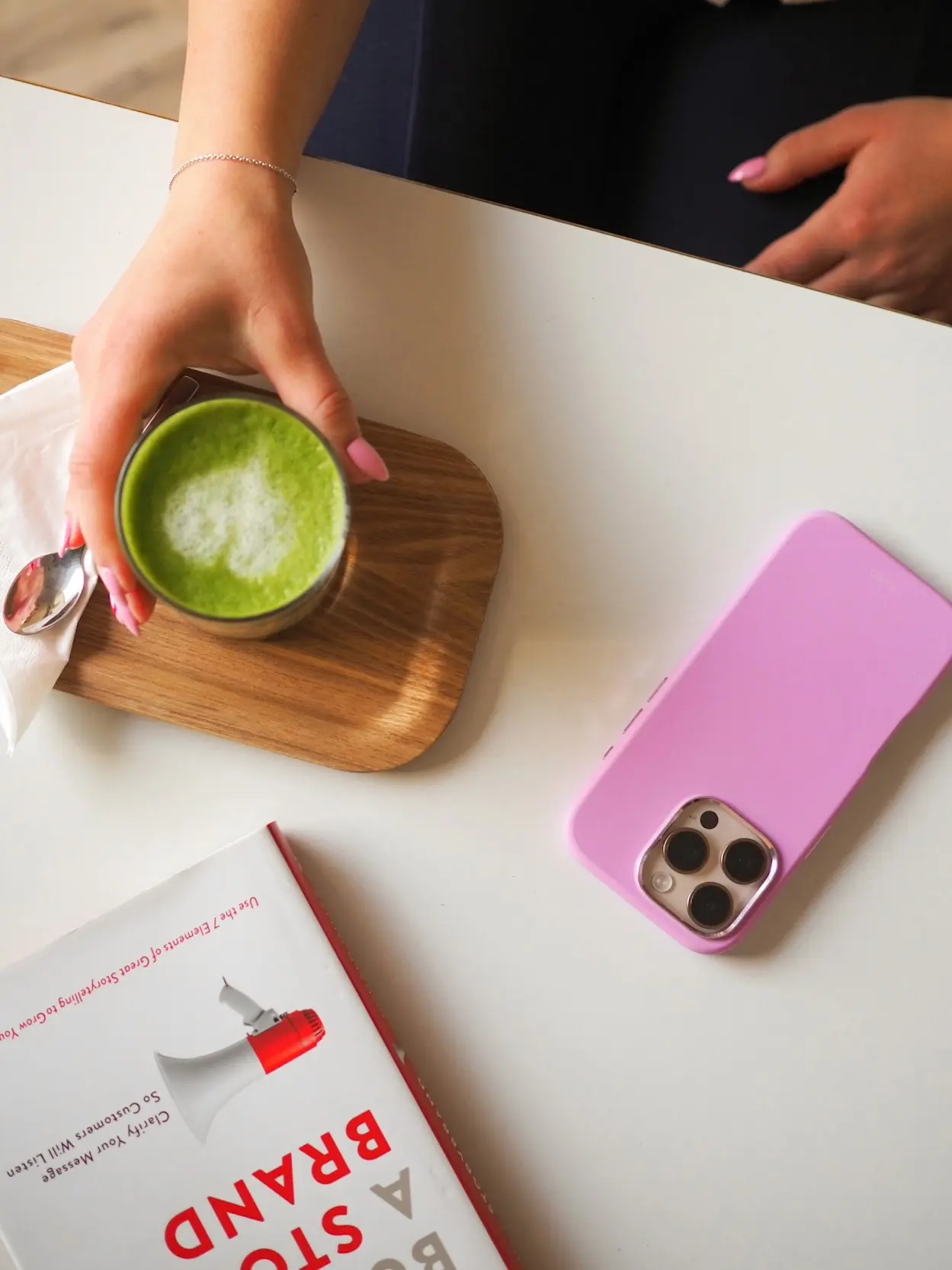 Käsi pitää matcha-lattea puisella tarjottimella, vieressä vaaleanpunainen puhelin ja kirja.