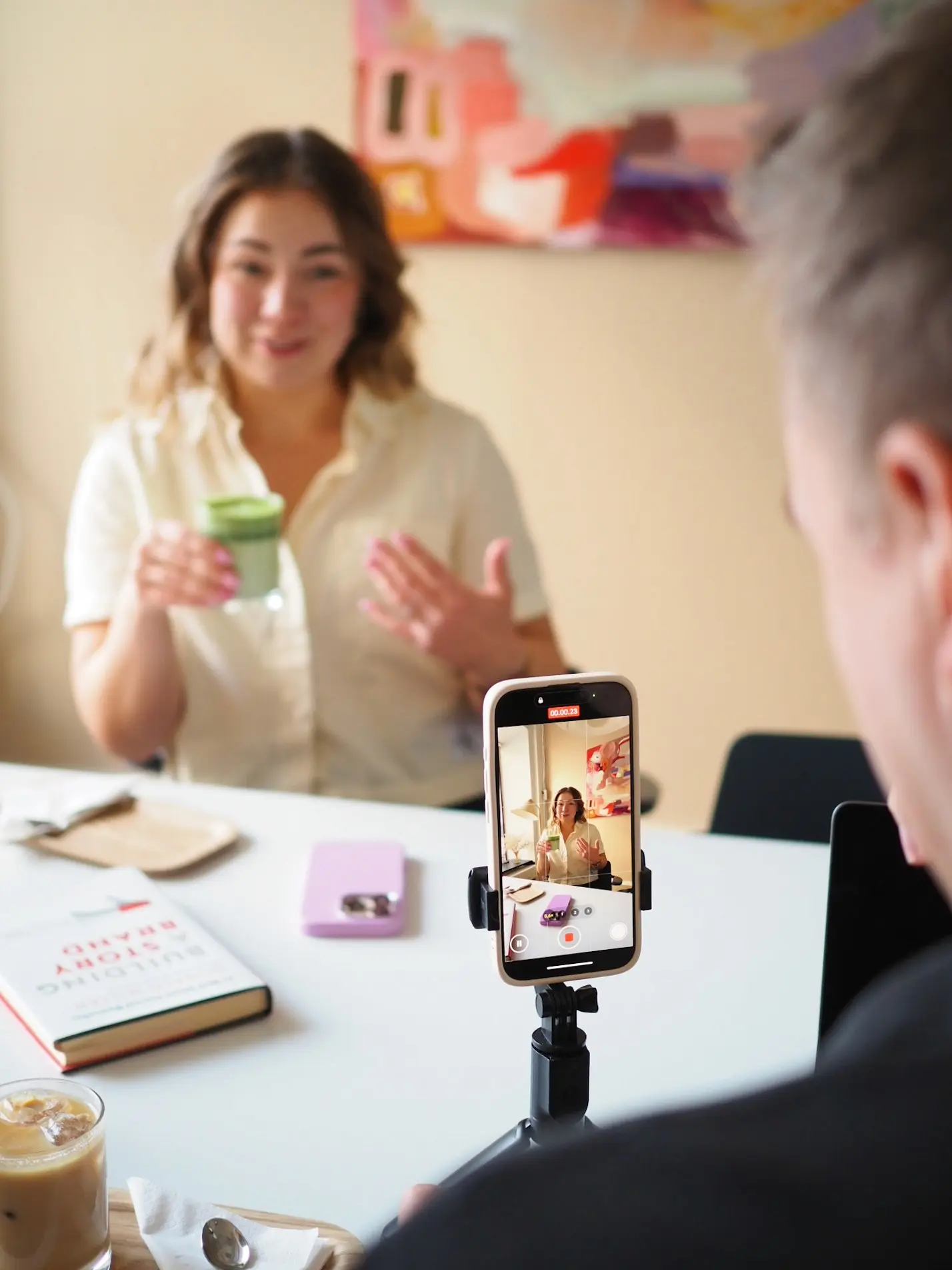 A person sits at a table talking to the camera while holding a green drink, filmed on a phone mounted on a tripod. On the table are a book, a phone and a drink. A short-form video is being shot.