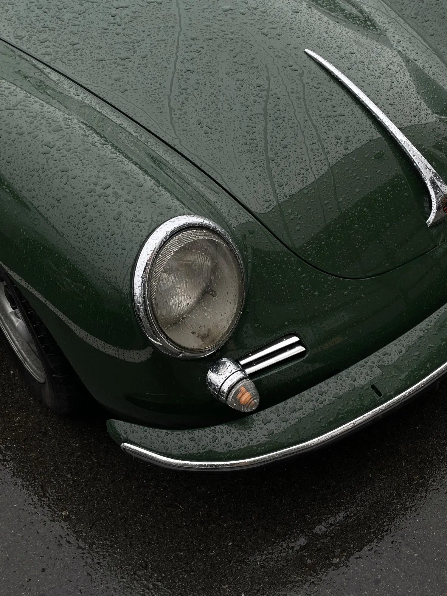 The front corner of a green classic car after rain, with water droplets on the surface and chrome details such as the headlight and indicator catching the light.