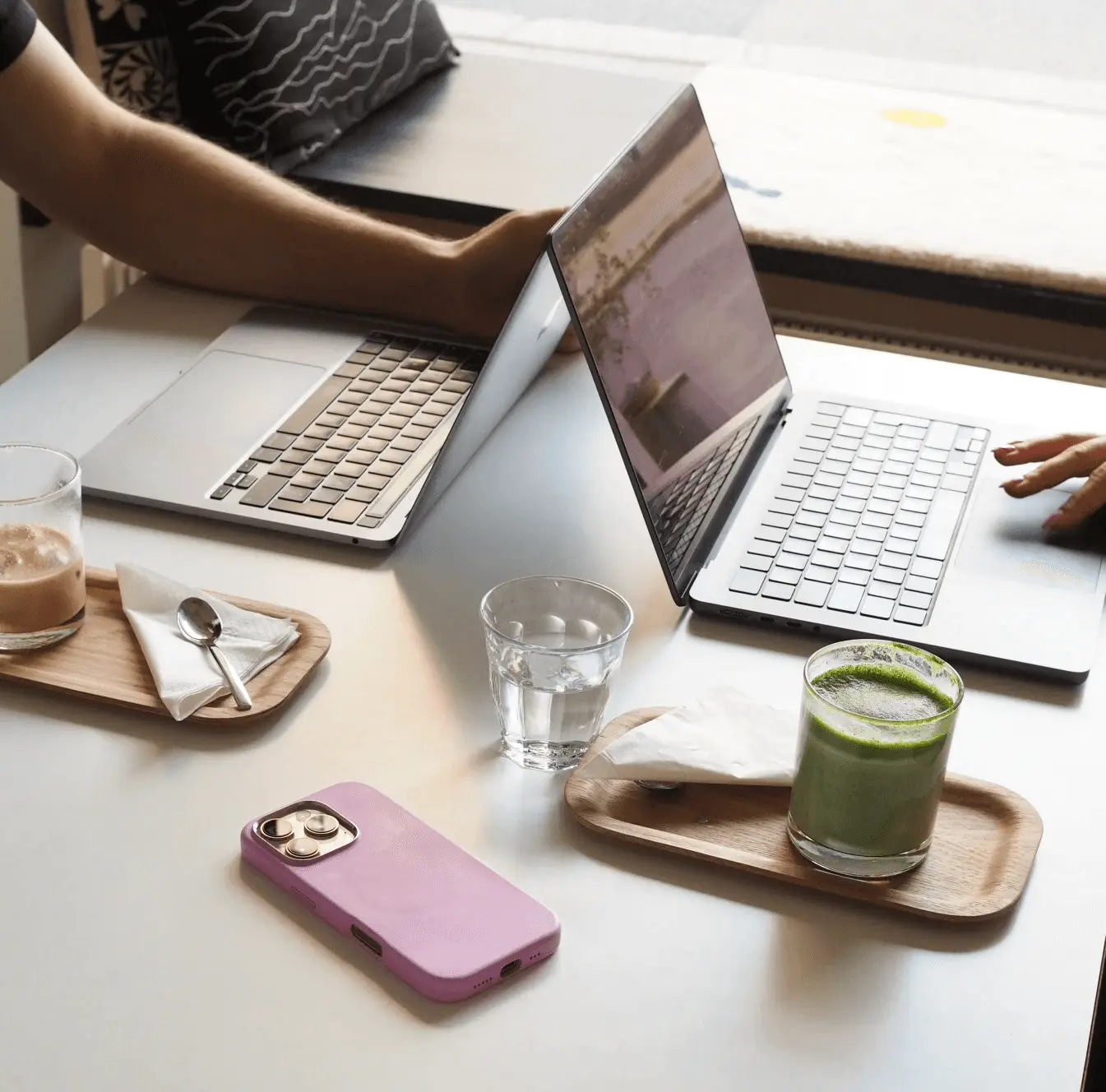 Two people work on laptops at a cafe table with a green drink, a coffee, a glass of water and a lilac phone. They are planning or working on a short-form video.