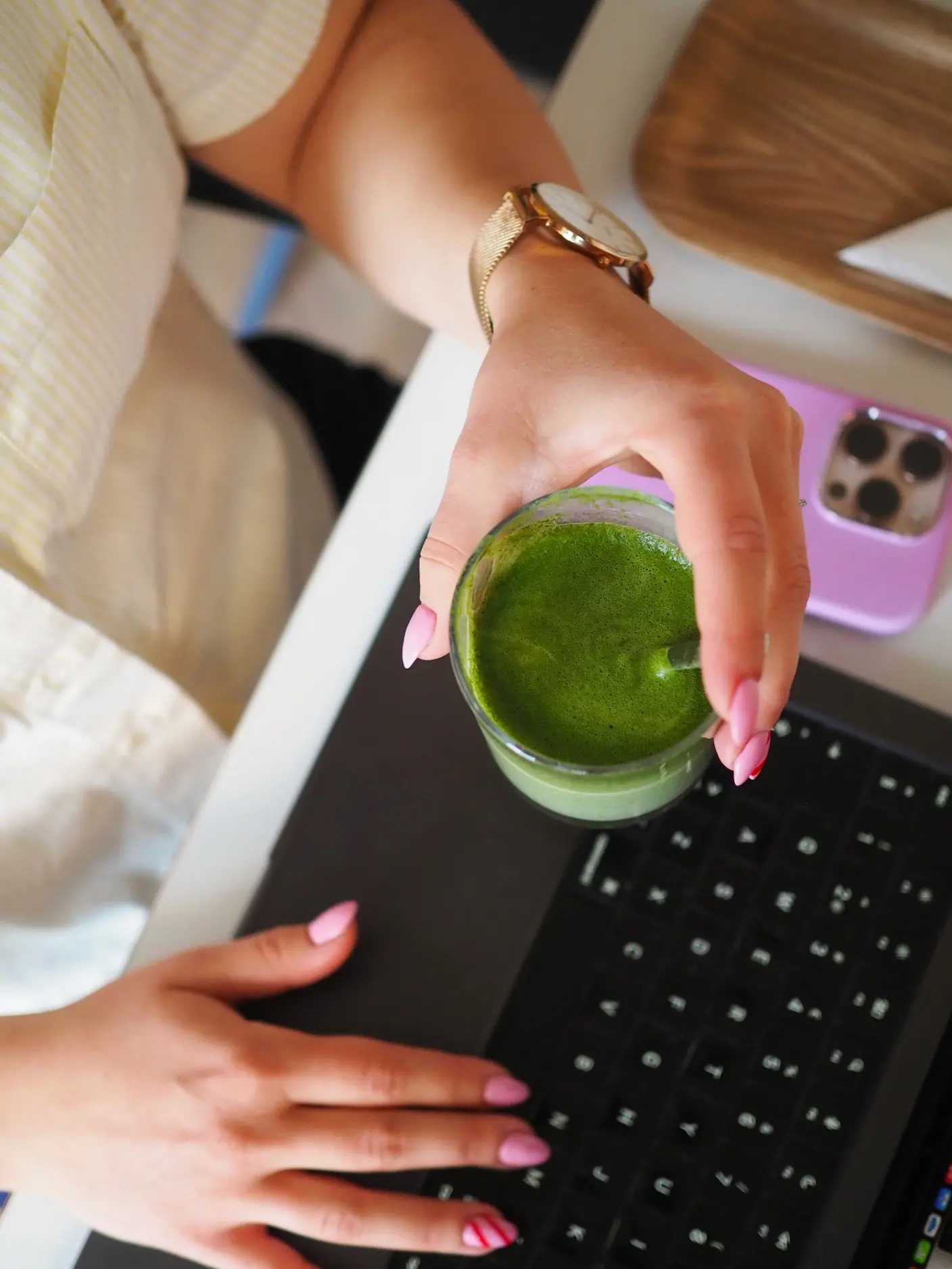 A person holds a green drink above a black laptop; a lilac phone and part of a white shirt are also visible on the table.