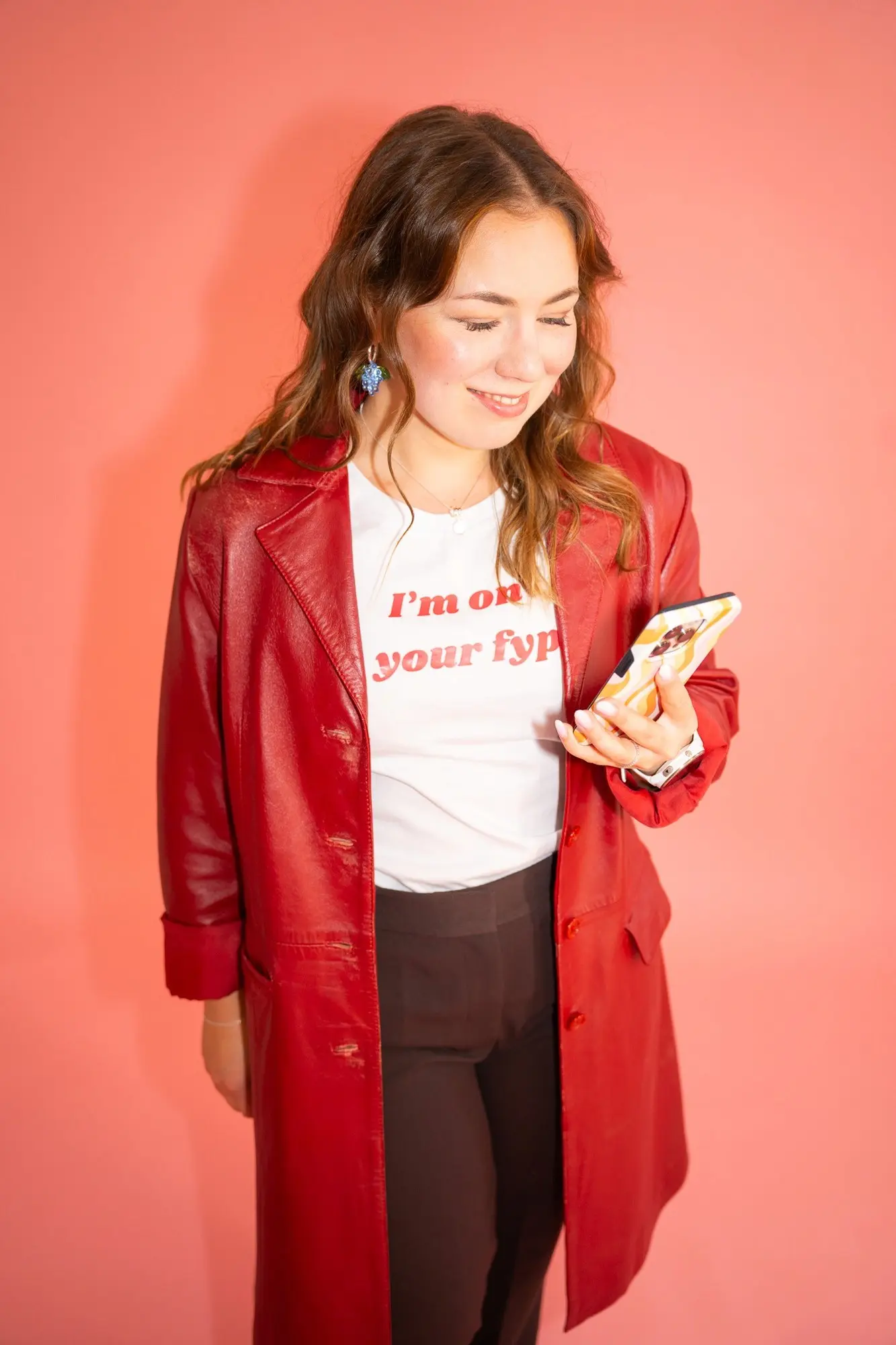 MAU Agency CEO Saana Nieminen stands in front of a pink background, wearing a red leather jacket and a white T-shirt that reads ‘I’m on your fyp’. She is smiling and looking at her phone.